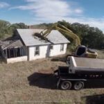 Excavator demolishing old house near dump truck